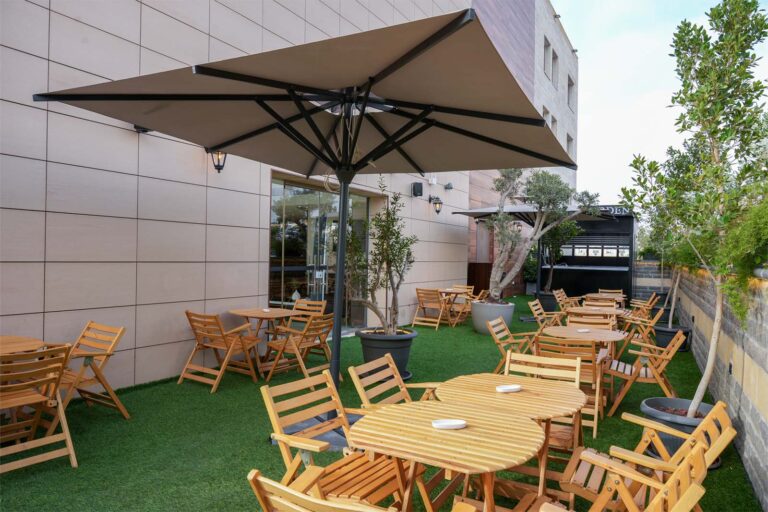 Outdoor café seating area with round wooden tables and matching folding chairs under large beige umbrellas on artificial grass.