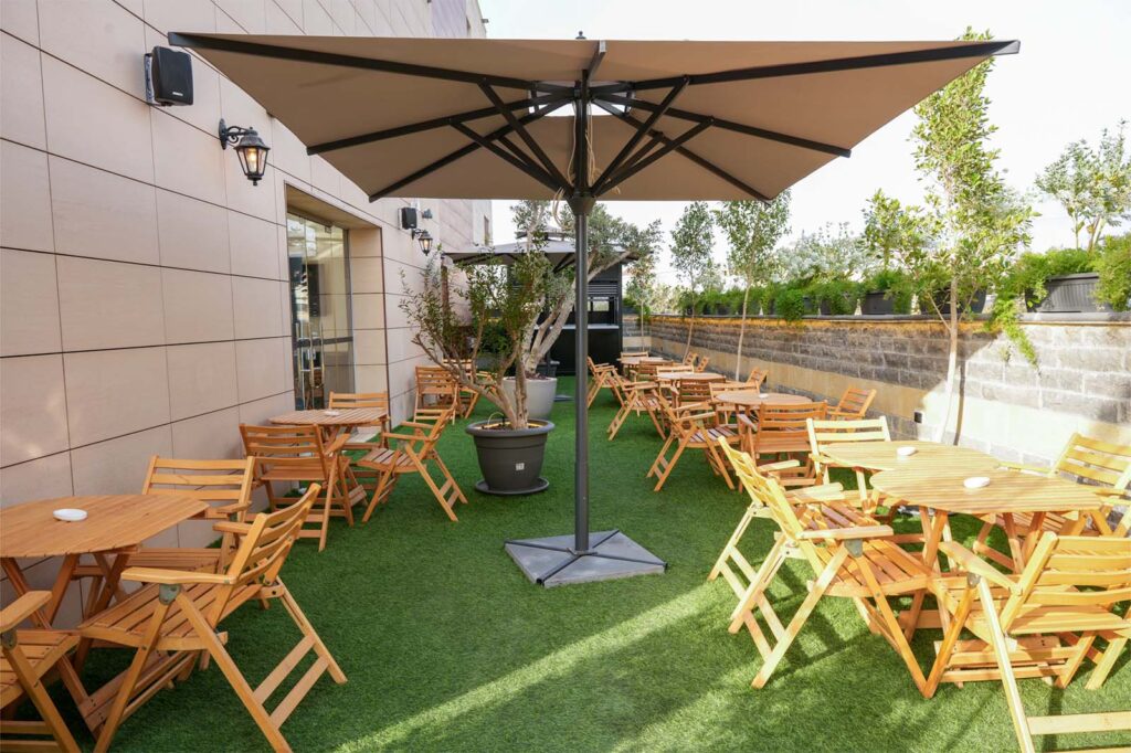 Outdoor dining patio with wooden tables and chairs under a large beige umbrella on artificial grass.