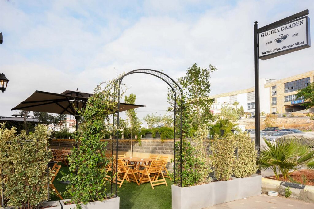 Outdoor café seating area with wooden tables and chairs under umbrellas, surrounded by greenery and planters.