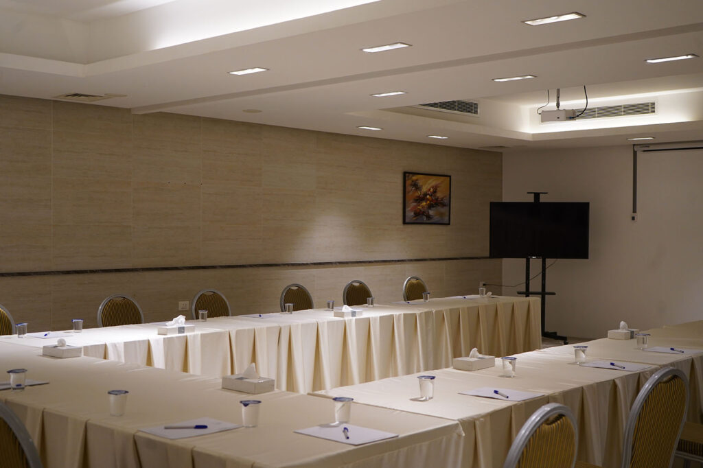 Conference room prepared for a meeting with U-shaped beige-draped tables, cups, notepads, and a large screen on a stand at the front.