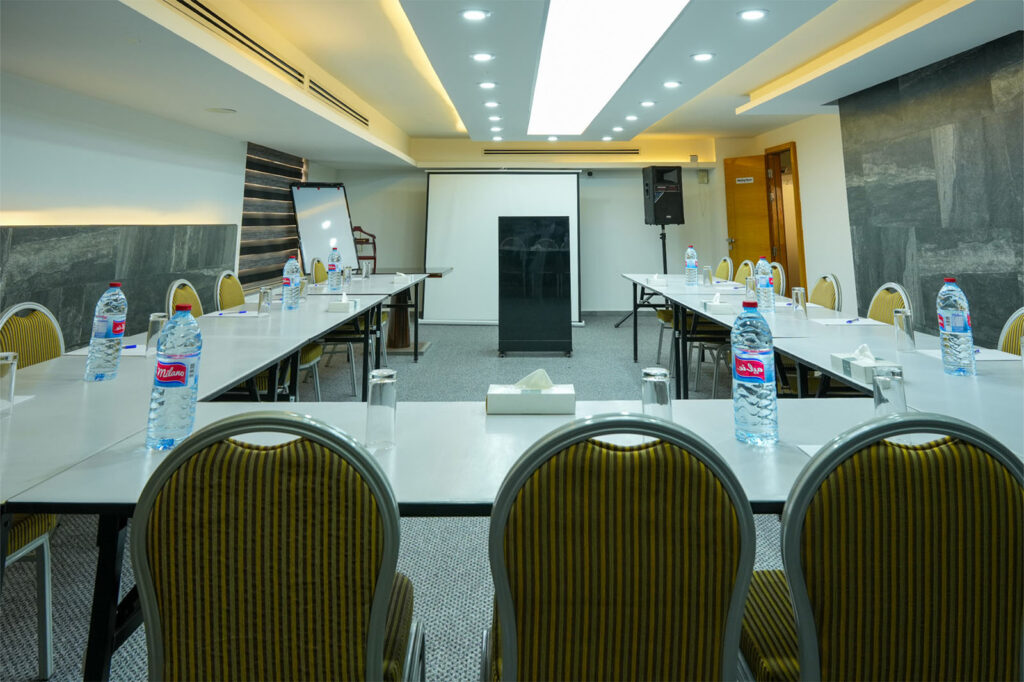 Conference room setup with U-shaped tables, bottled water, tissue boxes, and a projector screen at the front.