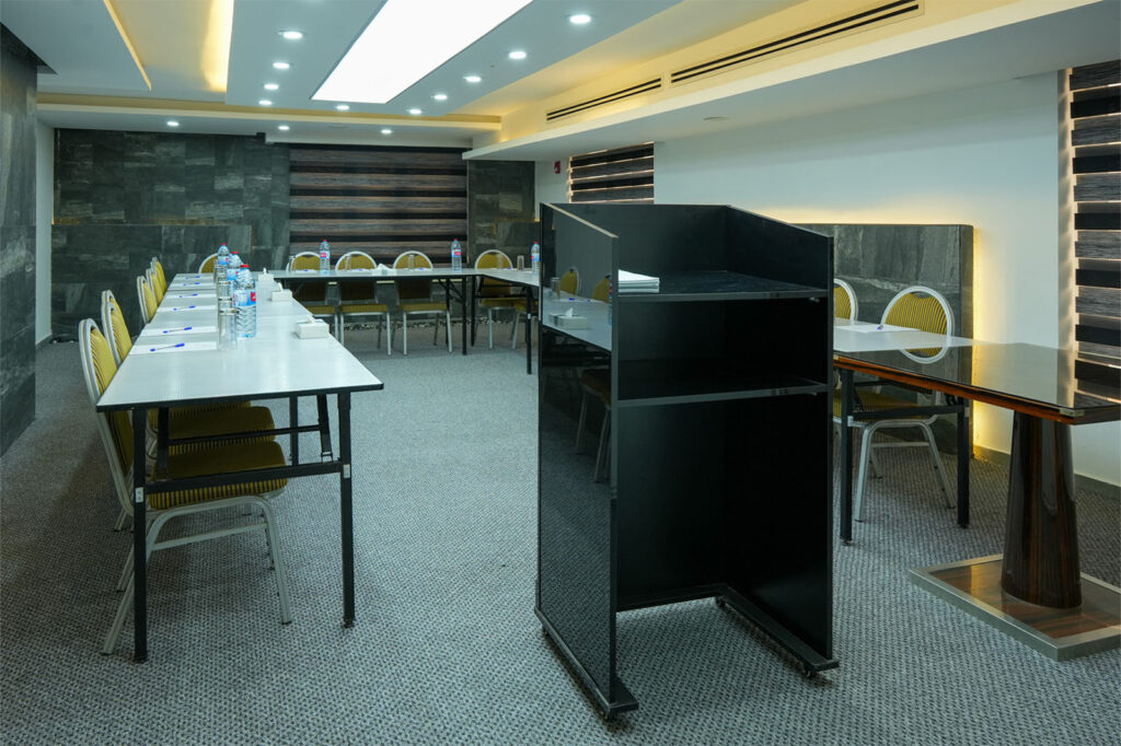 Conference room arranged for a meeting: long tables with yellow chairs, water bottles, and a black podium in the foreground.